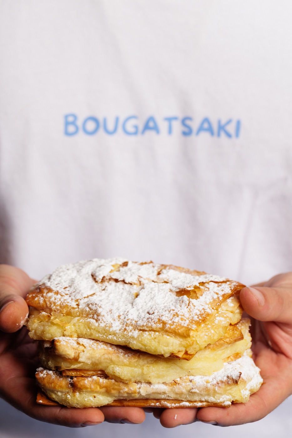 A stack of golden, hand-dusted bougatsa held in two hands, wearing a Bougatsaki apron